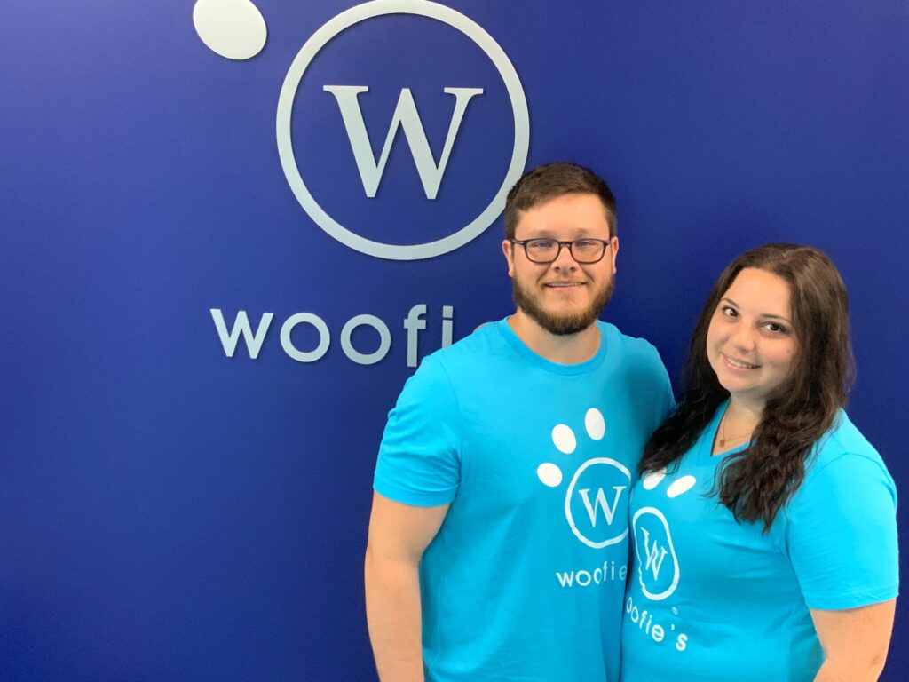 A man and women smiling in front of Woofie's logo on a blue wall